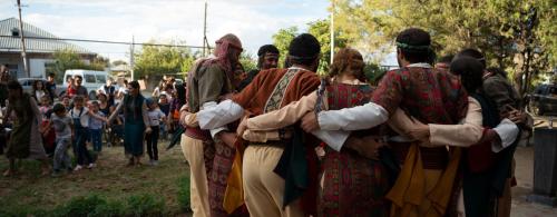 To the right, a group of adult men and women dance in a circle, hands clasped around each other's backs. To the left, young children also dance hand in hand.