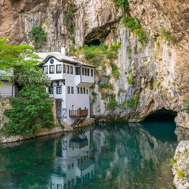 Three-story white building with dark gray trim situated at the edge of a turquoise waterway and the bottom of a sheer rock cliff.