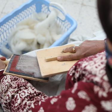 Close-up over a person's shoulder as they card strands of grayish white wool. A basket of uncarded wool is at their feet.
