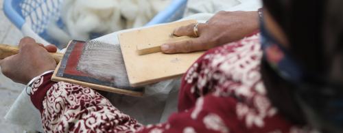Close-up over a person's shoulder as they card strands of grayish white wool. A basket of uncarded wool is at their feet.