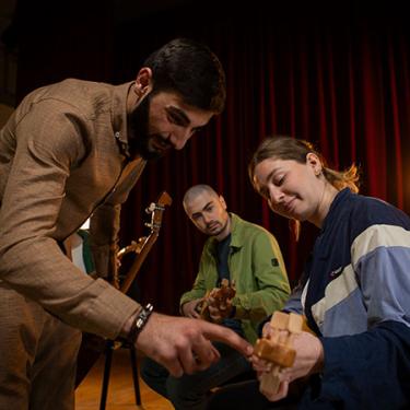 One person plays a string instrument while two others watch, and one points to part of the fretboard.