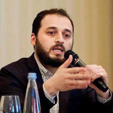 A person with short dark hair and beard wearing a suit jacket speaks into a microphone and gestures with one hand.