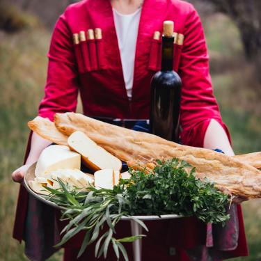 A person, head out of frame, holds a silver platter of breed, cheese, leafy greens, and a wine bottle.