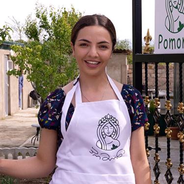 A young woman wearing a white apron and dark blouse smiles.