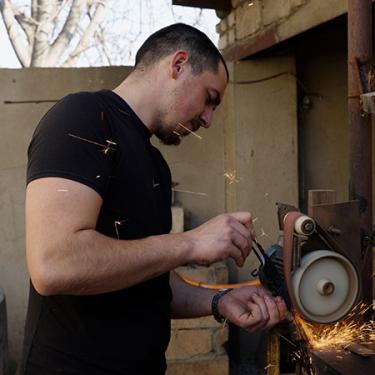 The young man in the Nike shirt holds the blade of a knife to a mechanical sharpening tool in an outdoor workshop.