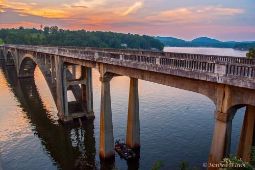 An arched, concrete bridge over a river, under a pink sunset. Photo credit in lower right: Matthew H Irvin.