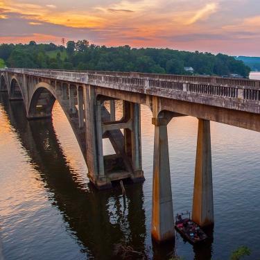 An arched, concrete bridge over a river, under a pink sunset.
