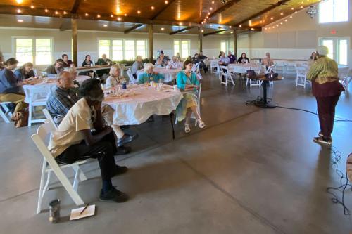 Groups of people sit at tables during a presentation by one woman.