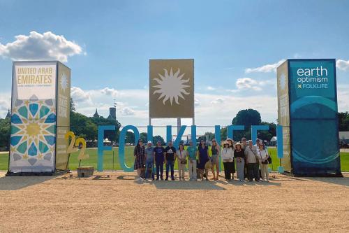 Fifteen people pose in front of a yellow and turquoise marquee that reads 22 FOLKLIFE! between tall banners for program on United Arab Emirates and Earth Optimism, and underneath a banner with the Smithsonian sunburst logo, on the National Mall.