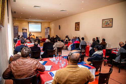 A group of people seated at tables face the front of the room, where a woman is giving a slide presentation. Many of them wear headphones for translation devices.