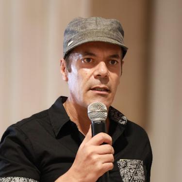 A man wearing a black dress shirt and cap speaks into a microphone