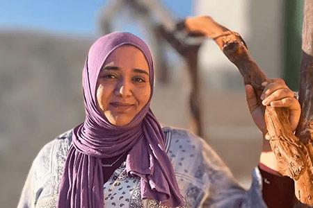 A woman smiles, wearing a purple headscarf and holding onto a branch.
