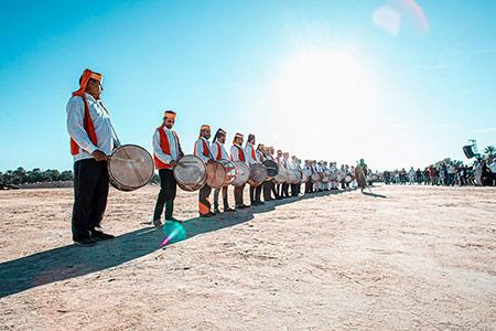 A line of drummers wearing red vests and white shirts stand in the dessert on a clear, blue-skied day.