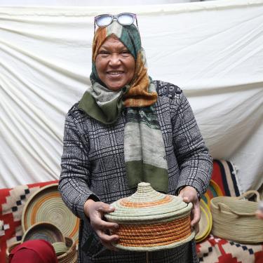 A woman wearing a long-sleeved printed black and white dress and colorful headscarf holds up a circular tan and orange basket woven from natural materials.