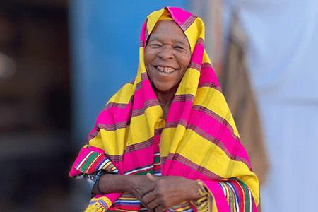 A women smiles, wearing a headscarf with bright pink and yellow stripes.