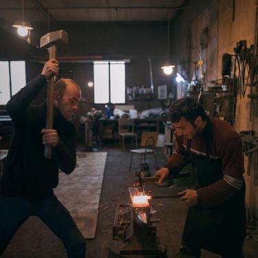 Two blacksmiths at work in a shop, pounding a glowing-red metal rod.