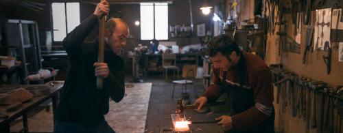 Two blacksmiths at work in a shop, pounding a glowing-red metal rod.