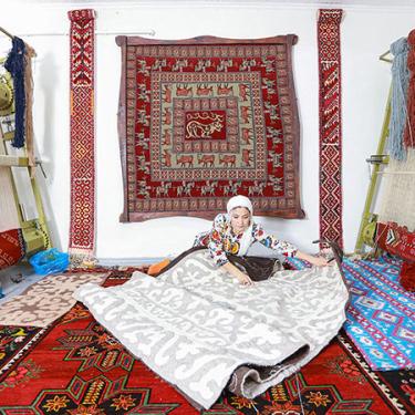 A woman sits on the ground, unrolling a woven gray and white rug. More rugs sit under her and hang behind her, and on either side are two large looms with rugs in progress.