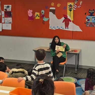 An adult sits in a classroom while reading a book. Children sit on a rug and listen.