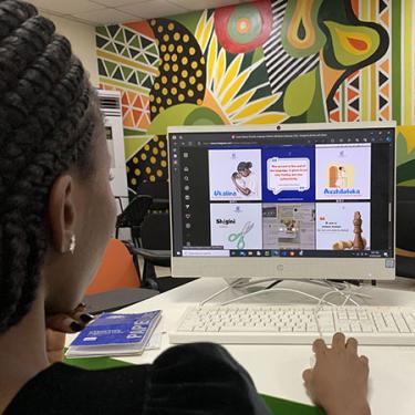A woman at a computer looks at images of educational language content with a colorful mural in the background.