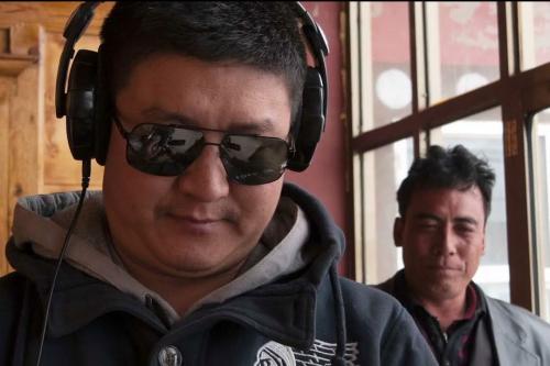 two men looking at a screen out of view. One wears sunglasses and headphones