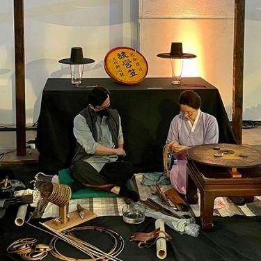 An older woman and a young man sit next to each other on a floor. The woman in front holds a flat wooden disk in the shape of a hat to bend horsehair around.
