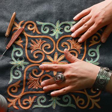 Close-up on two hands touching raised needlework in turquoise and orange on a dark gray textile background.
