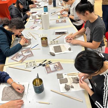  In a dim, gray room, five men sit among huge, detailed Mongolian masks. One man uses a tool to work on one of the masks, while the other four watch.