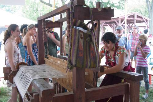 A woman works on a large loom in front of a small crowd of people outdoors.