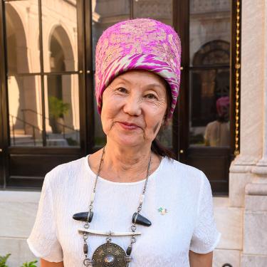 A woman wearing a white top, beaded necklace, and pink head covering stands outdoors, smiling.
