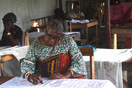 A woman applies dye or to create a pattern on fabric. Other women at work stations behind her also work on textiles.