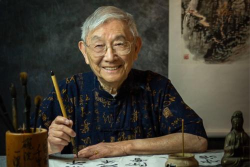 A man with gray hair and glasses sits at a desk and smiles. He is holding a calligraphy pen, and in front of him on the desk sits intricate Chinese calligraphy.