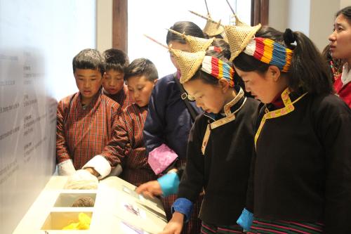 A group of young students, the girls in black traditional jackets and woven conical hats and the boys in red plaid shirts, inspect a museum exhibit of textile fibers.
