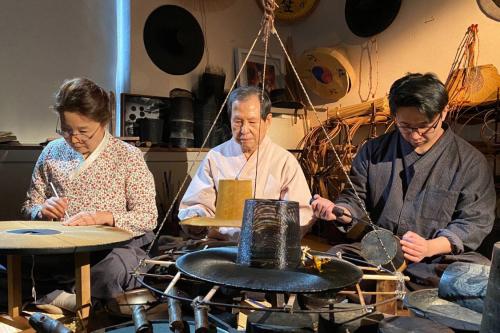 Three people— two men and a woman— sitting side by side in a room making horsehair hats, surrounded by other hats they've made.