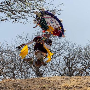 A man in feathered blue, white, and red headdress dances outdoors on a hill. Barren trees are in the background. 