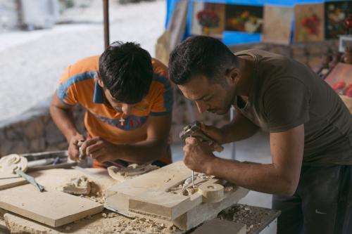 two men bent over, holding chisels carve pieces of wood on a table.