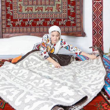 A woman sits on the ground, unrolling a woven gray and white rug. 