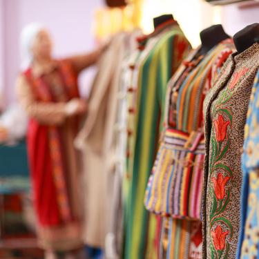  In the foreground, a lineup of mannequins each with colorful embroidered garments. At the end of the line, out of focus, a woman dresses a mannequin.