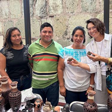 Four people pose in front of a table full of ceramic vases and ornaments. The person on the right holds up two tiny clay  pots. 