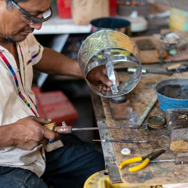 A man wearing safety glasses sits at a work table, holding a screwdriver-like tool in one hand and a cylindrical silver tin art piece in the other. 