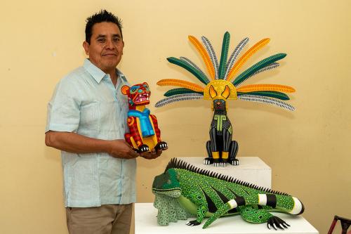 A man poses holding a red and orange sculpture of a lion or other teeth-bearing large cat, wearing a bright blue scarf, next to culptures of a green iguana and a black cat with green and orange feather headdress.