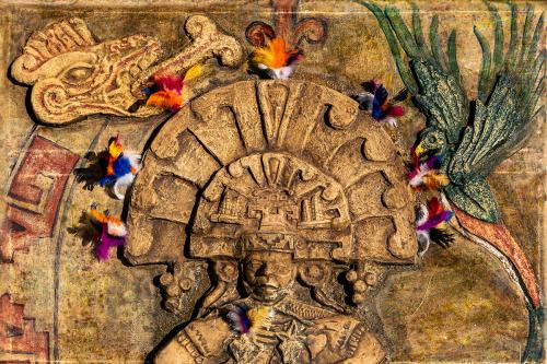 Stone relief art of a person wearing a semicircular headdress, adorned with brightly colored feathers.