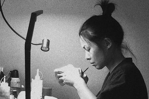 A woman with her hair in a bun sits at a desk, repairing a piece of pottery.