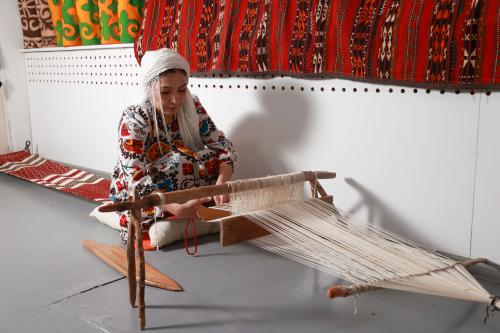 A woman wearing a white headdress and floral tunic sits on the ground, weaving. She looks down, concentrating on the loom.