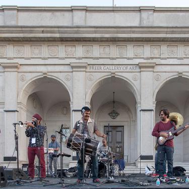 A band performs on an outdoor stage. The lead singer is playing a large drum slung over his shoulder.