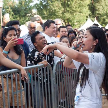 A woman raps at an outdoor festival, with a crowd of people singing along on the other side of a small gate.