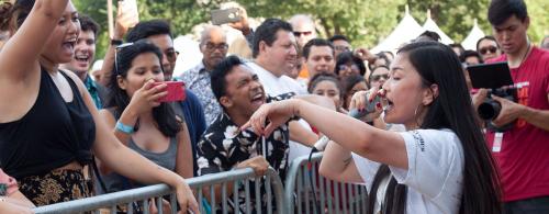 A woman raps at an outdoor festival, with a crowd of people singing along on the other side of a small gate.