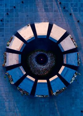 From overhead, a circular memorial structure with 12 trapezoidal columns surrounding a central ring. Thousands of flowers and flower wreaths are placed around a lit flame in the center.