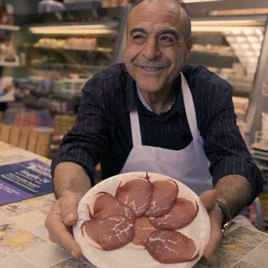 Smiling and standing behind the counter of his sandwich shop, a man holds out a plate of Basturma, a cured beef.
