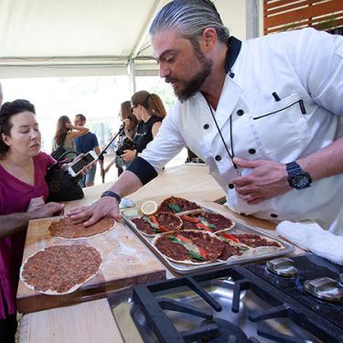 A man with grey hair in a white chef’s coat leans over his cooking station, checking on the flatbread he is preparing. A woman to his left looks on.